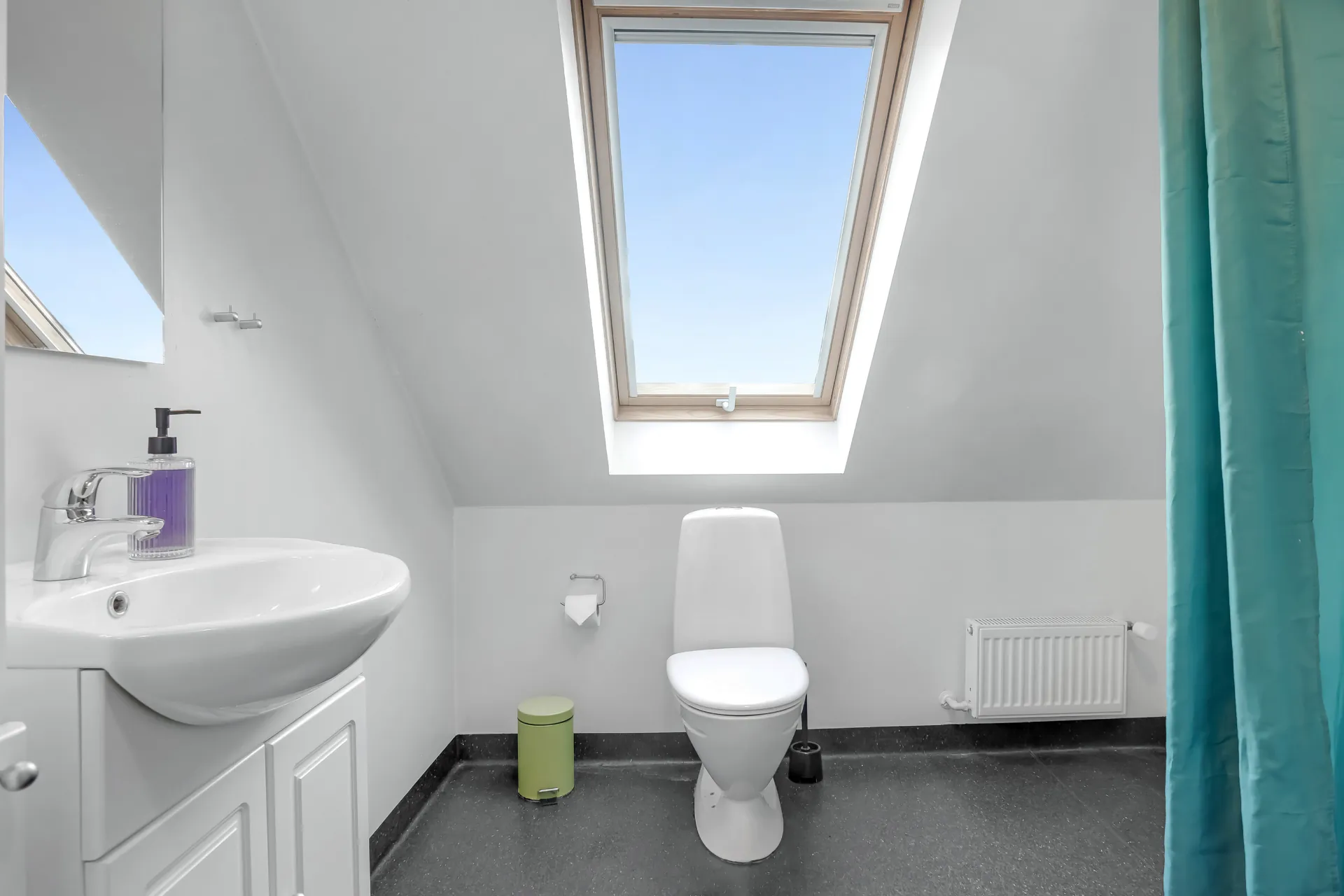 Håndvask med spejl og toilet under et stort ovenlys, og et brusegardin i turkis