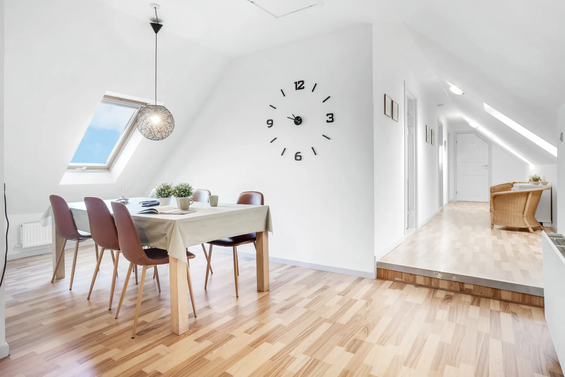 Spisebord med stole under skråvæggen, kuglelampe over bordet og et stort vægur på væggen